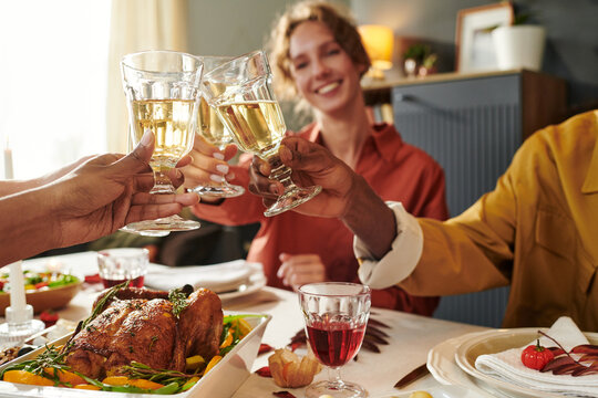 Multiethnic group of young adults clinking wine glasses while sitting around dining table with roasted chicken and salad, smiling and celebrating together during Thanksgiving meal