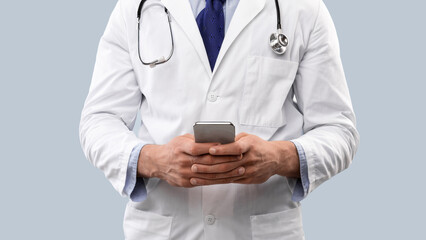 Cropped Shot Of Doctor Man Using Smartphone With Medical Application Standing In Studio Over Blue Background. Unrecognizable Professional Physician Posing Browsing Internet On Cellphone. Panorama