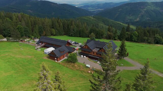Aerial view of wooden buildings with dark roofs nestled amidst lush green fields, surrounded by dense forest and rolling hills, Jablunkov, Moravian-Silesian Region, Czechia.