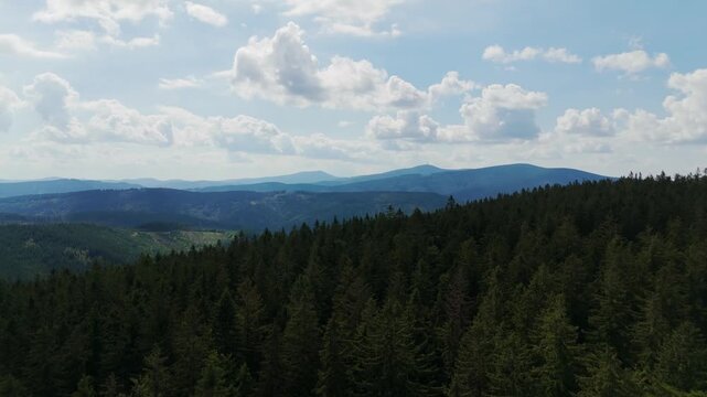 Aerial view of a dense forest with rolling mountains in the distance under a bright blue sky dotted with fluffy white clouds, Jablunkov, Moravian-Silesian Region, Czechia.