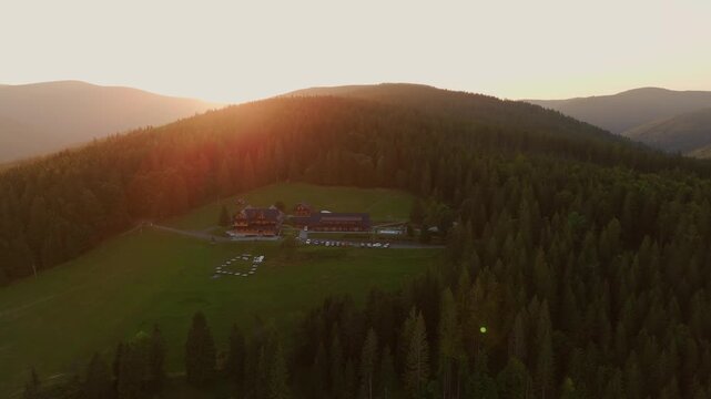 Aerial view of a mountain lodge, nestled amidst lush green hills and dense forests, as the sun casts a golden glow, Jablunkov, Moravian-Silesian Region, Czechia.