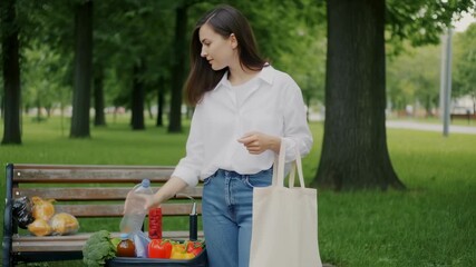 A woman in a park sorts groceries on a bench, holding a tote bag. Produce is visible - Powered by Adobe