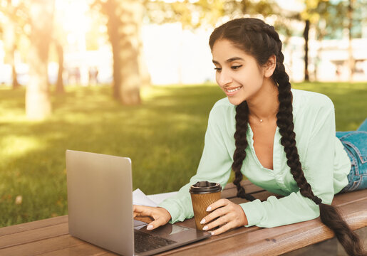 Rest at campus. Happy indian college student girl relaxing outdoors with coffee and laptop, lying on bench in park, smiling at computer screen, free space