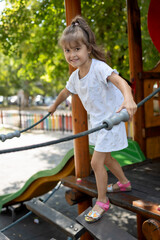 Adorable little girl with a happy smile playing on a wooden rope bridge at the playground. Cheerful child enjoying a sunny summer day of active fun and adventure outdoors.