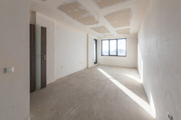 Interior of an empty room under construction in a new apartment. Unfinished space with raw concrete walls, floor, and a large window. Renovation and real estate concept.