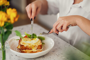 Person cutting into cheesy baked lasagna with fried egg, served with arugula on white plate. Casual dining scene with yellow flowers and stylish manicure.