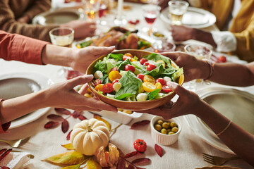 Multiethnic group of adults sitting around table sharing large bowl of fresh salad, hands of...
