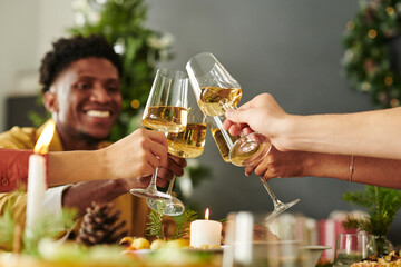 Group of young adults clinking wine glasses during Christmas celebration, multiethnic friends smiling and toasting together at decorated holiday table
