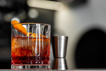 Close-up of amber cocktail with clear ice cube and orange peel garnish in faceted glass on reflective surface, bar background with jigger, elegant lighting and mixology vibe