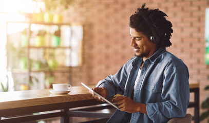 Cheerful young african american curly man in wireless headphones look at tablet, sit at table with cup of coffee in cafe interior. Study remote with modern gadget, funny video and surfing in internet