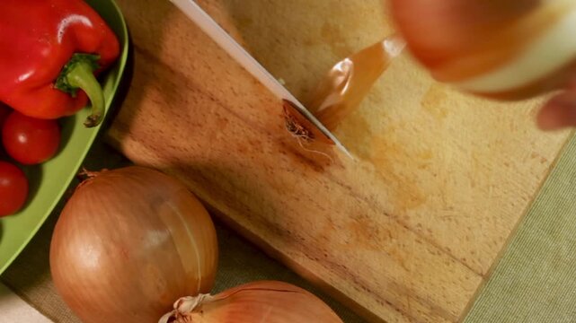 Hand peels golden onions on wooden board with knife, carefully removing skin and cutting off ends. Peeling onions before cooking. Concept of home cooking and clean produce.