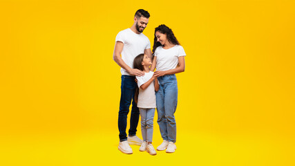 Arabic Parents And Little Daughter Posing Hugging And Holding Hands Smiling To Each Other Standing Together On Yellow Studio Background. Vertical Shot Of Happy Middle-Eastern Family Of Three