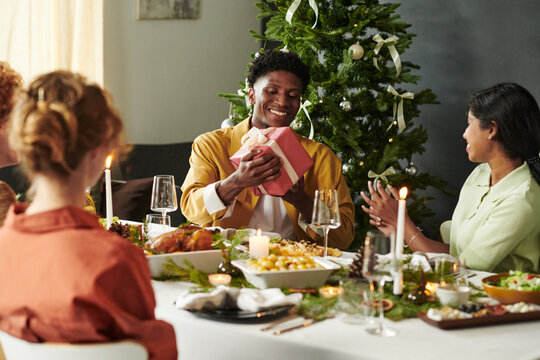 Young Black man smiling while receiving wrapped gift from young Asian woman at Christmas dinner table with multiethnic friends, Christmas tree decorated in background, celebrating holiday together - Powered by Adobe