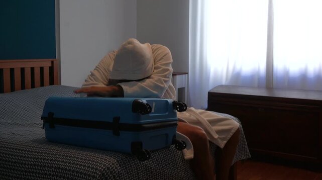 Desperate man in depression at home in bedroom cries desperately near his suitcase after returning and finishing a trip - difficulty returning home to normality and work after a long trip abroad