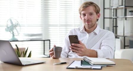 Fototapeta premium Man with cup of drink and smartphone at wooden table indoors