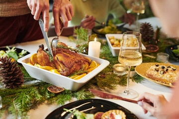Caucasian adult carving roasted chicken at Christmas dinner table surrounded by multiethnic adults enjoying holiday meal with wine, candles, pinecones and seasonal decorations