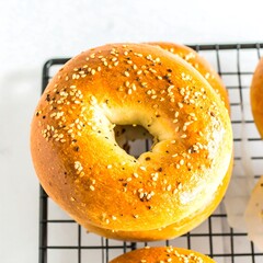 A close-up shot features a freshly baked bagel, coated with sesame seeds and other seasonings, on a black wire rack with a light background