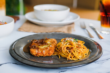 
A fusion meal featuring crispy chicken schnitzel with gravy and a side of spaghetti bolognese, served on a dark plate on a marble table in a restaurant