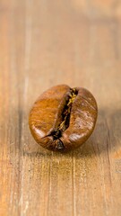 A close-up shot features a single, roasted coffee bean resting atop a textured, light-brown wooden surface. The bean?s dark-brown exterior reveals a slight crack