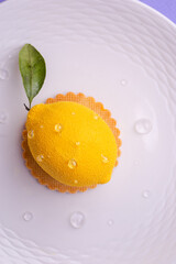 Overhead shot of a hyper-realistic lemon-shaped dessert: textured surface, dew-like droplets, and green leaf on a ridged white plate.