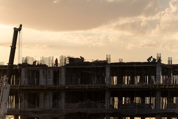 Silhouettes of construction workers on a building under construction against a beautiful, cloudy sky during the golden hour. A crane is visible on the left side of the frame.