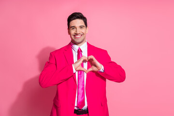 Cheerful young gentleman in vibrant pink suit making heart sign on pink background for celebration...