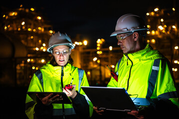 Team of construction engineers in uniforms and helmets examine project with tablet Industrial plant or oil refinery background at night