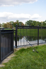 Fishing rod leaning against metal fence near riverbank in city park. Urban outdoor scene with...