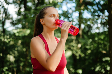 A focused young woman in a red athletic top is taking a drink from a bright pink water bottle while...