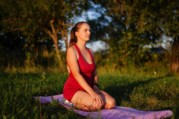 
A calm woman in a red athletic suit is sitting kneeling (Vajrasana) on a purple yoga mat in a sunny, grassy park during golden hour, meditating or resting after a workout.