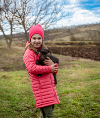 Smiling girl in red jacket and beanie holding black baby goat on farm. Rural lifestyle, childhood, animal care countryside setting