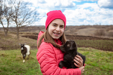 Smiling girl in red jacket and beanie holding black baby goat on farm. Rural lifestyle, childhood, animal care countryside setting