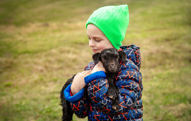Joyful boy holding black baby goat on farm. Rural lifestyle, childhood, and animal care in countryside setting. Happy childhood