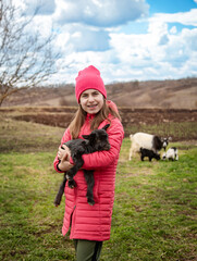 Cute girl in red jacket and beanie holding black baby goat on farm. Rural lifestyle, childhood, animal care countryside setting