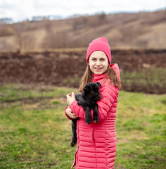 Cute girl in red jacket and beanie holding black baby goat on farm. Rural lifestyle, childhood, animal care countryside setting