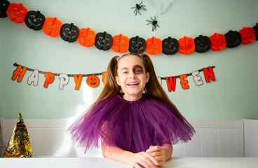 Smiling girl purple tulle costume with Halloween makeup, sitting decorated room with pumpkins, garlands, spiders, and witch hat.