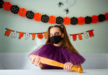 Girl dressed in purple costume with face makeup and black face mask holds wooden bat in room decorated for Halloween.