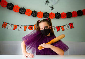 Girl dressed in purple costume with face makeup and black face mask holds wooden bat in room decorated for Halloween.