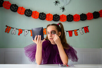 Girl wearing purple costume applies spooky makeup using mirror, surrounded by festive Halloween decorations