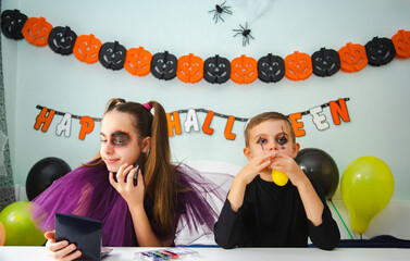 Two kids getting ready for Halloween. One girl applies makeup while boy inflates balloon.