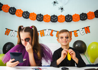 Two kids getting ready for Halloween. One girl applies makeup while boy inflates balloon.