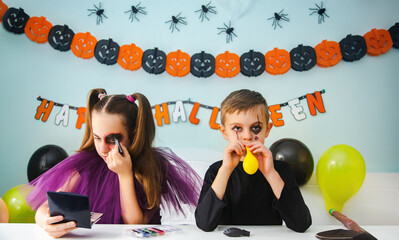 Two kids getting ready for Halloween. One girl applies makeup while boy inflates balloon.