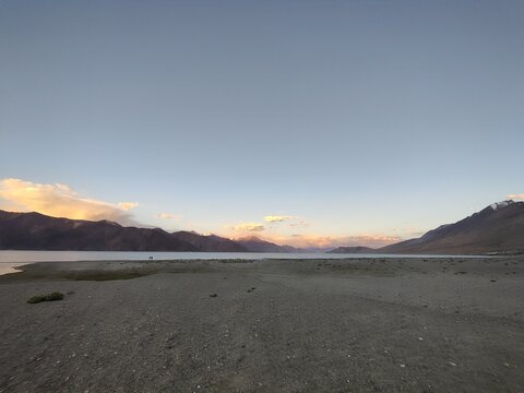 Peaceful mountain lake at sunset with calm water, clear sky, and distant hills glowing in soft evening light, showing natural beauty and quiet atmosphere.
