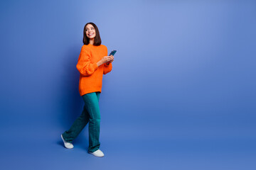 Smiling young woman in orange sweater holding smartphone against vibrant blue background, showcasing casual style