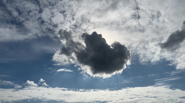 A low-angle view of a dynamic sky features a large, dark cloud in the center, partially blocking the sun, surrounded by bright white cumulus clouds and a vivid blue sky