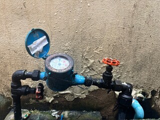 Close-up of an old, worn blue water meter and connecting pipes with rusty valves mounted against a rough, weathered stucco wall, highlighting aging utility infrastructure.
