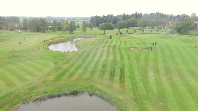 Aerial footage of a golf course with visible fairways, sand bunkers, ponds, and golfers playing under cloudy skies in a scenic countryside landscape.
