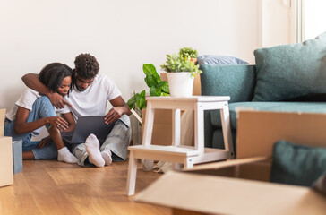 Couple sitting on the floor surrounded by moving boxes, using a laptop in their new home. Cozy atmosphere with plants, couch, and natural light symbolizing a fresh start and togetherness