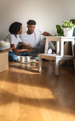 African American couple sitting on the floor surrounded by boxes, drinking coffee and using a laptop to plan home furnishing and decoration after moving into their new apartment.
