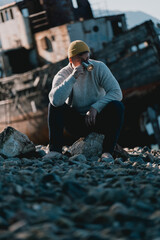 A man in a yellow beanie and gray sweater sits on a rock, drinking from a cup near a rusted...